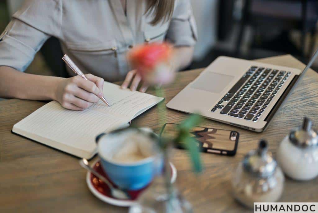 Person Writing On A Notebook Beside Macbook